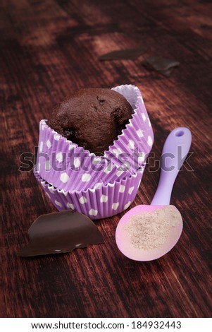 Chocolate cupcake in purple dotted paper baking form, chocolate bar and baking mixture on wooden background. Baking cupcakes rustic style.