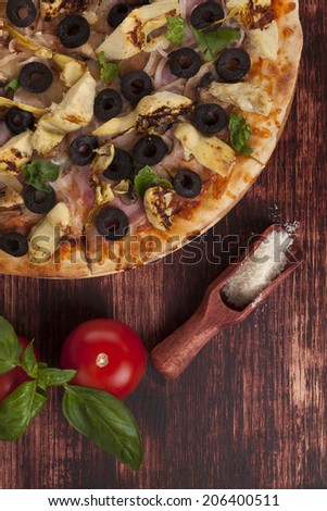 Rustic, country style pizza background. Pizza with black olives, ham and artichokes on wooden background with parmigiano cheese, tomatoes and basil.