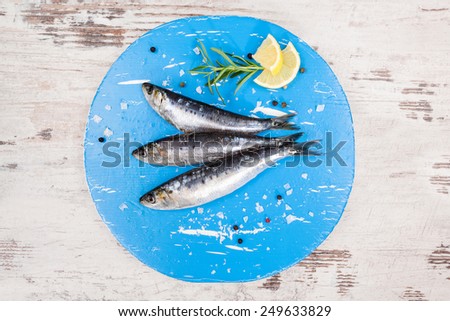 Three fresh anchovy fish on blue round kitchen board on white wooden table, top view. Culinary seafood concept. Delicious healthy eating.