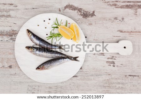 Three fresh anchovy fish on white round kitchen board on white wooden table, top view. Culinary seafood concept. Delicious healthy eating.