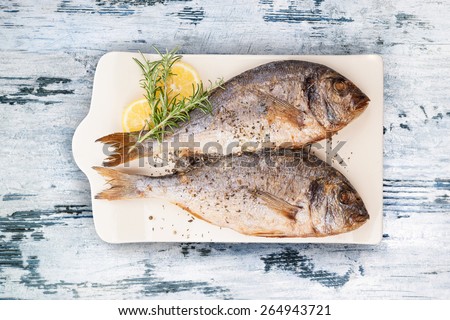 Delicious grilled sea bream fish on kitchen board with rosemary, lemon and colorful peppercorns on white textured wooden background. Culinary healthy cooking. Stock photo.