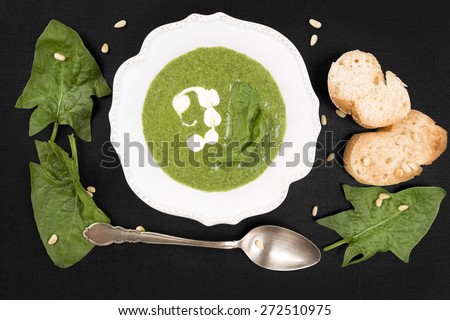 Delicious fresh spinach soup with fresh spinach leaves and bread in white vintage plate on black stone background, top view. Culinary soup eating.
