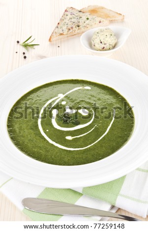 Spinach soup in white plate on wooden table, toast bread and herbal butter in bowl in background.