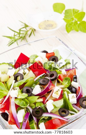 Greek salad in bowl close up, olive oil dressing and fresh herbs in background.