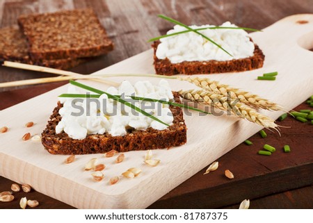 Black whole wheat bread slices with cottage cheese, fresh herbs and wheat on wooden cutting board. Healthy rustic eating background.