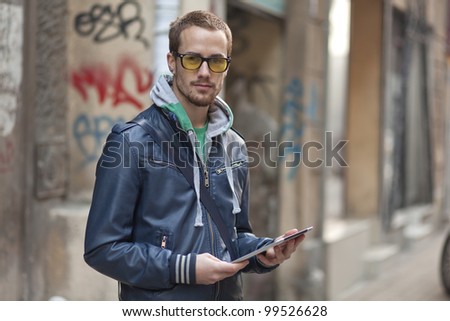 Young man with yellow glasses use tablet computer on street, public space. Blurred background