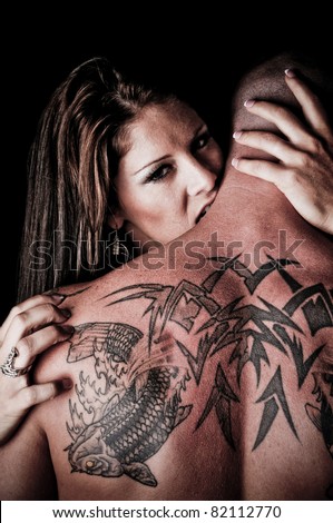 Beautiful Woman biting, hugging and looking over the shoulder of a man with tattoos
