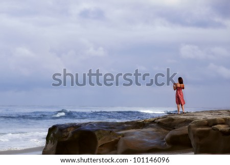 Woman in red vintage summer sun dress standing on a rock bluff along the sea coast watching surfers and enjoying the tropical California waves of the Pacific ocean