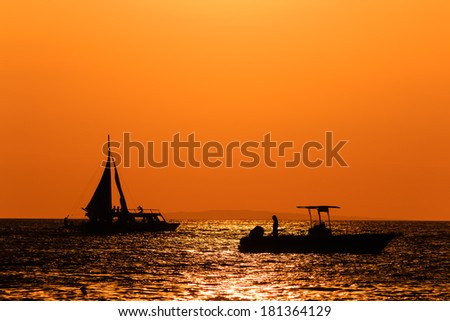 Silhouette of boats against an orange sunset sky on a calm ocean