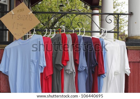 Red, white, blue and gray t-shirts hung up on a porch railing on hangers to sell as part of items at a yard sale. A sign on the left offers the t-shirts for a $1. each.