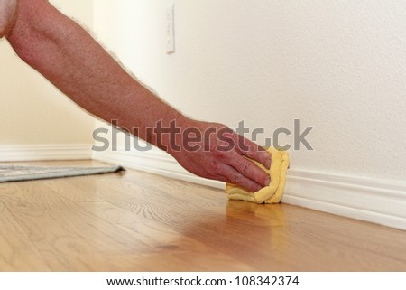 Male caucasian hand and arm seen wiping a folded yellow rag along the lower white wall trim near the wood floor to remove dust.