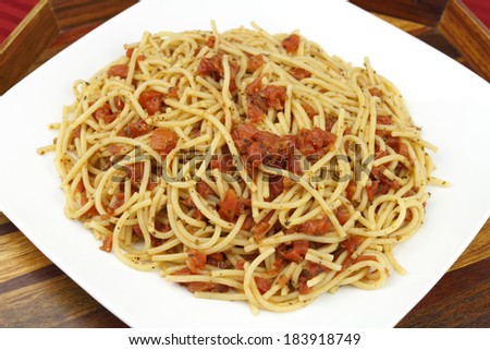 Delicious looking dinner of cooked spaghetti pasta noodles mixed with diced organic tomatoes and herbs served on a white plate with wooden serving tray on a red tablecloth.