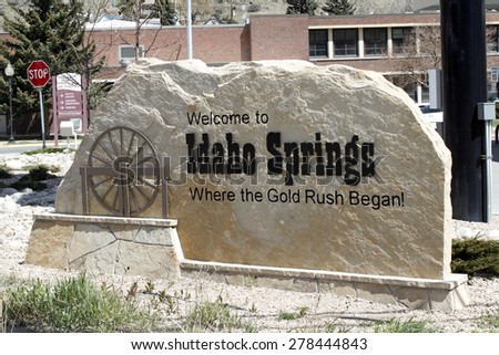 Idaho Springs, CO, USA - April 23, 2014: Large tan stone that has written on and carved into it, Welcome to Idaho Springs, Where the Gold Rush Began! The sign has a brown sculpture of a water wheel