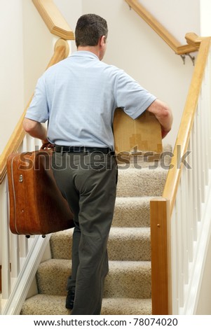 Man dressed in business casual clothes walking up stairs with suitcase in one hand and box under right arm.