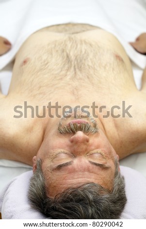 Mature man lying face up on a white sheet covered professional massage table waiting for a massage to relax his sore muscles and to feel better.