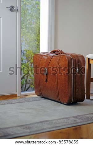 One brown suitcase sitting on the entrance rug inside the front door of a home in the daytime waiting to be taken on some travel.