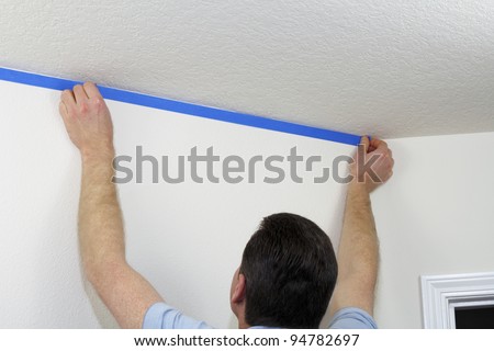 Man preparing to paint ceiling by masking off the wall beneath it with blue painter\'s tape.