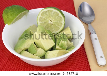 Avocado pieces in a white bowl with lime juice and lime wedges on the side of the bowl. A spoon is near by on a yellow napkin and everything is on a red placemat.