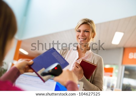 Portrait of young female college student returning book to library, with librarian scanning barcode