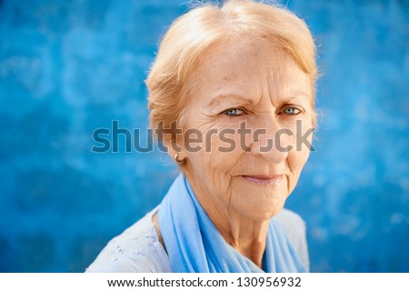 Senior people portrait, happy old blonde woman in blu clothes smiling and looking at camera against blue wall