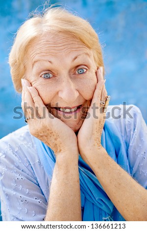 Old blonde woman portrait, lady in elegant clothes with hands on face for joy and surprise on blue background