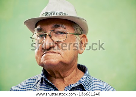 people and emotions, portrait of serious senior hispanic man with glasses and hat looking at camera against green background