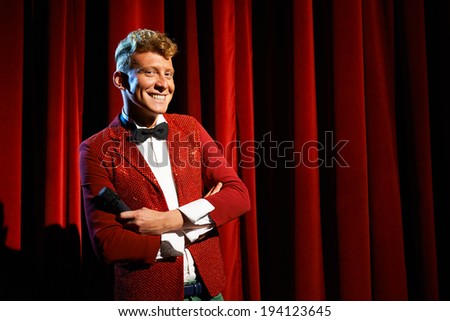 Arts and entertainment in theatre with funny man working as anchorman, standing with arms crossed against red curtains with microphone