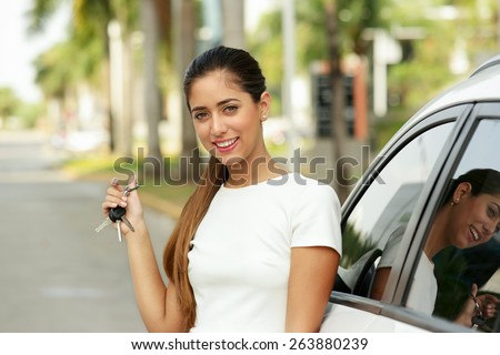 Young adult hispanic woman holding keys of new white car, leaning on auto door. She smiles and looks proud at camera, showing the keys. Waist up portrait