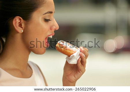 Close up of young hungry business woman with mouth open, doing breakfast eating a delicious pink donut from fast food on street. Side view, copy space