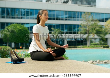 Young stressed hispanic business woman doing yoga outside office building, sitting in lotus position with hands on knees in the street. Concept of long working hours and need of stress free break