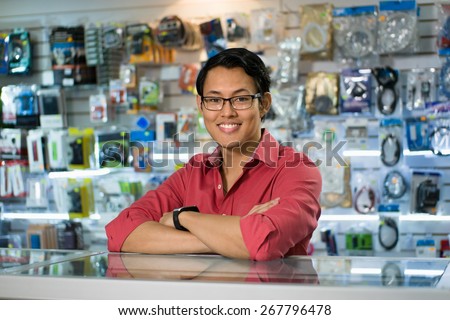 Portrait of young chinese man working as clerk in computer and technology store, smiling at camera and leaning on desk in shop