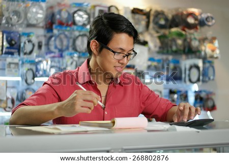 Young asian entrepreneur owning a computer shop and checking bills and invoice with calculator, doing budget and reviewing expenses.
