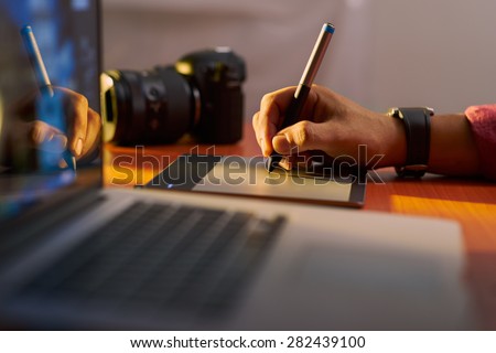 Photographer drawing and retouching image on laptop computer, using a digital tablet and stylus pen. Closeup of man\'s hand with dslr camera in background. Copy space in foreground