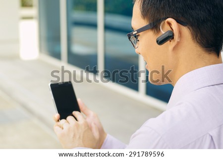 Young chinese businessman typing phone number on smartphone and talking with bluetooth headset device