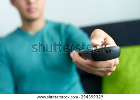 Asian young adult man watching TV and changing channel with remote control. Narrow focus on buttons and hand in foreground, cropped view.