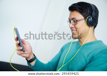 Chinese man relaxes on sofa and watches podcast on mobile phone, listening with green big earphones. Copy space