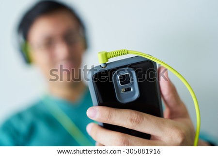 Chinese man watching podcast on mobile phone, listening with green big earphones. Copy space and selective focus on cable