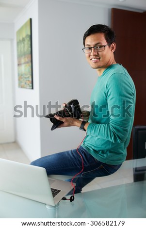 Chinese man connecting digital dslr camera to laptop computer via USB cable, watching photos and videos on PC. The guy sits on table in living room at home and smiles at camera