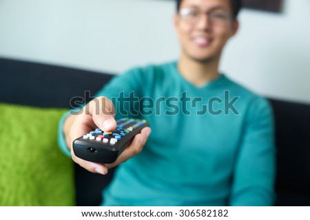 Asian young adult man watching TV and changing channel with remote control. Narrow focus on buttons and hand in foreground