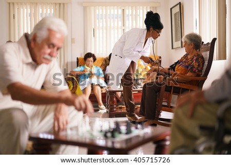 Old people in geriatric hospice: Black doctor visiting an aged patient, measuring blood pressure of a senior woman. Group of retired men in foreground playing chess.