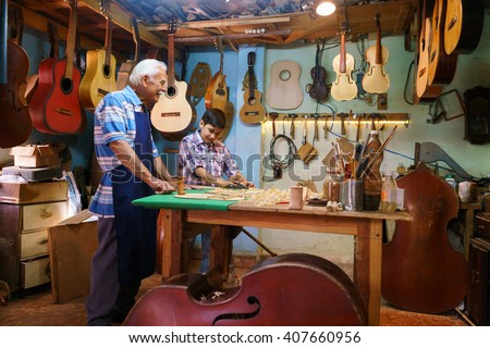 Small family business and traditions: old grandpa with grandson in lute maker shop. The senior artisan teaches to the boy how to chisel wood to make a music instrument