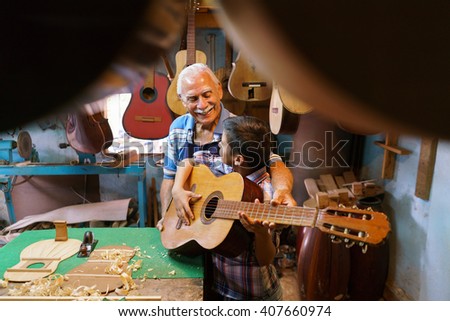 Small family business and traditions: old grandpa with grandson in lute maker shop. The senior artisan gives teaches how to play classic guitar to the boy, who plays his first musical notes