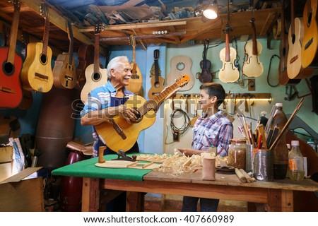 Small family business and traditions: old grandpa with grandson in lute maker shop. The senior artisan gives teaches how to play classic guitar to the boy, who looks carefully at the instrument