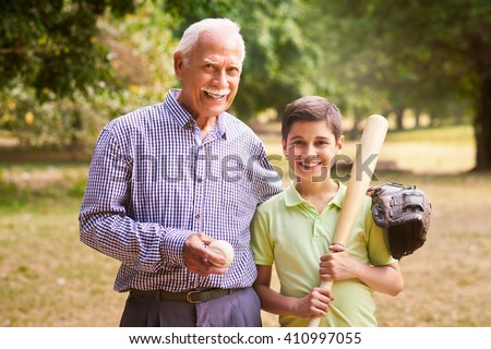 Grandpa spending time with grandson: Portrait of senior man playing baseball with his grandchild in park. The old man embraces the young kid holding the bat, smiling and looking at camera
