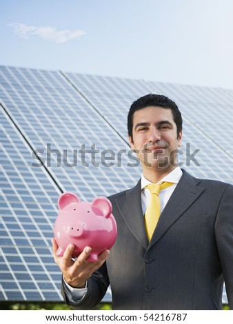 Portrait of mid adult italian male engineer holding piggy bank in solar power station, smiling at camera. Vertical shape, front view. Copy space
