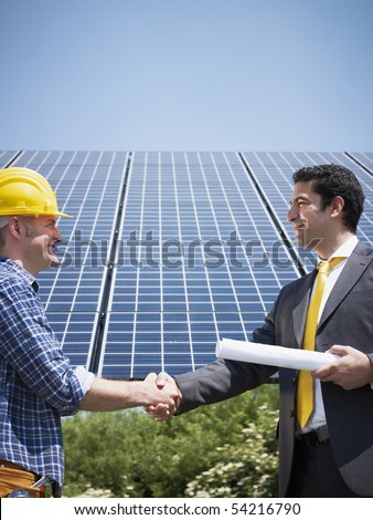 Portrait of mid adult italian male engineer holding blueprints and shaking hands to manual worker in solar power station. Vertical shape, side view. Copy space