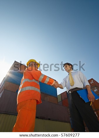Mid adult businessman holding clipboard and shaking hands to manual worker near cargo containers. Vertical shape, low angle view. Copy space