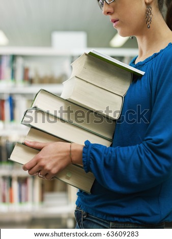 female college student carrying stack of books in library. Vertical shape, side view, mid section, copy space. Selective focus on face