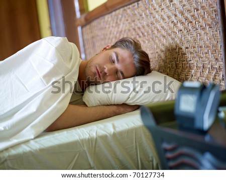 serene caucasian adult man lying in bed with alarm clock in foreground. Horizontal shape, waist up, front view