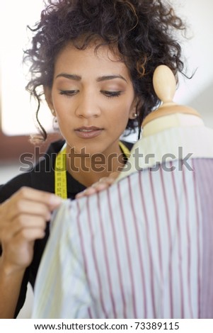 Young hispanic female dressmaker adjusting clothes on tailoring mannequin and smiling. Vertical shape, head and shoulders